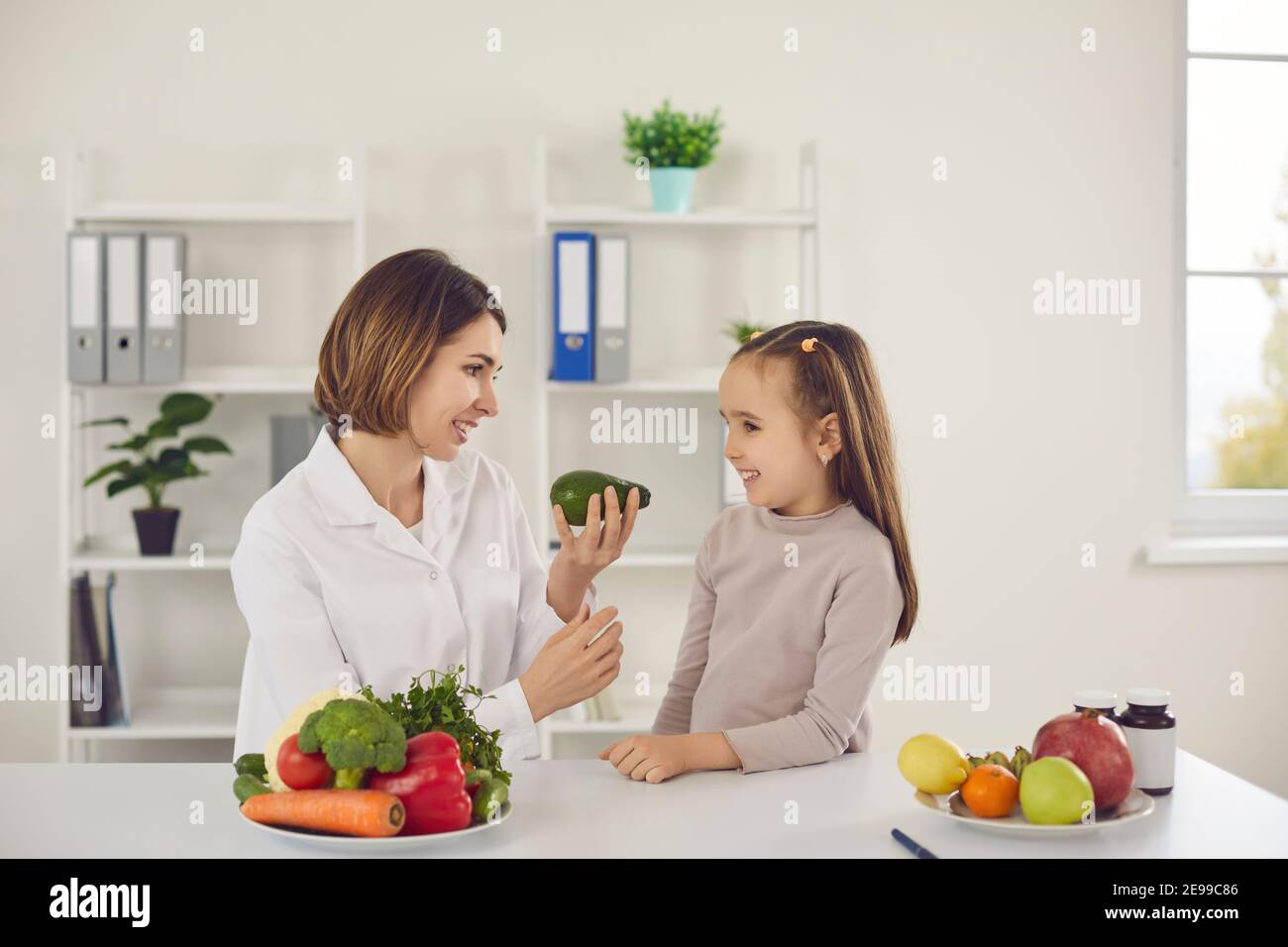 Nutritionist tells a little girl about the benefits of avocados while sitting at a table in office. Stock Photo