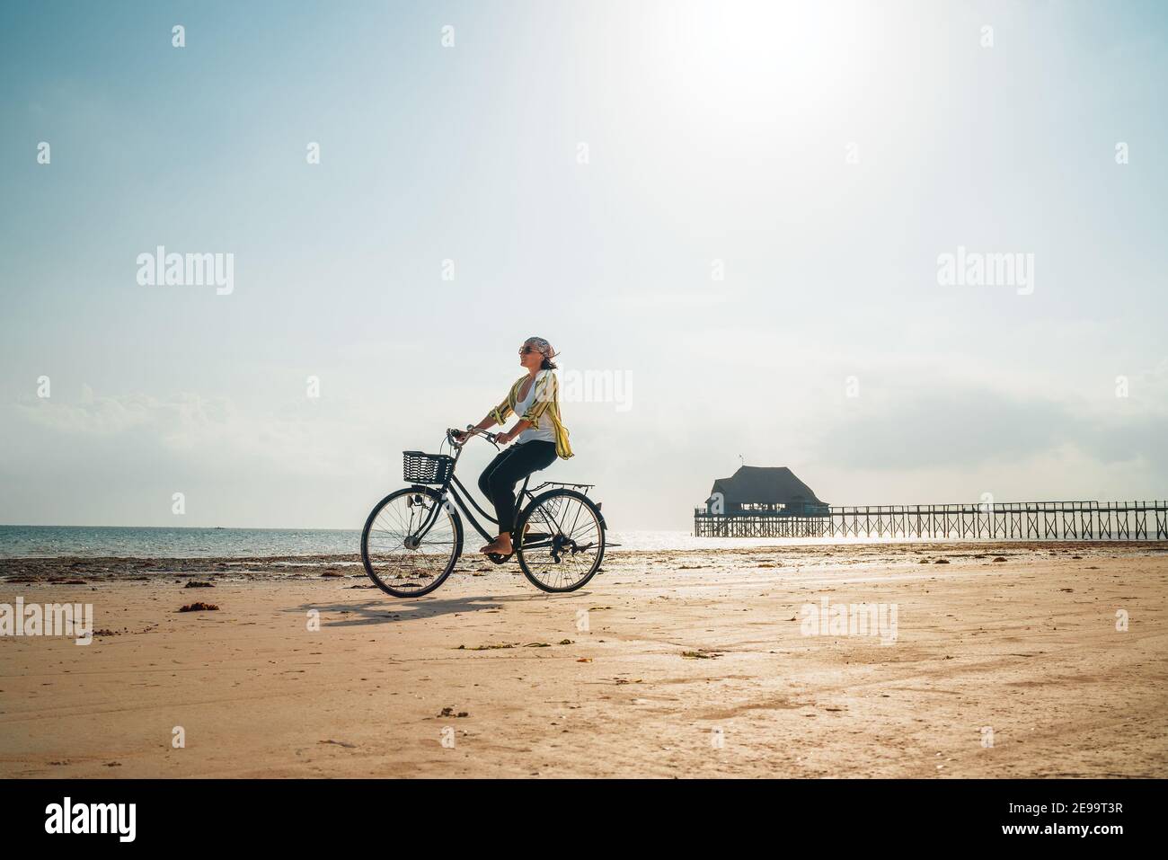 Young woman dressed light summer clothes riding old vintage bicycle with front basket on the lonely low tide ocean white sand coast on Kiwengwa beach Stock Photo