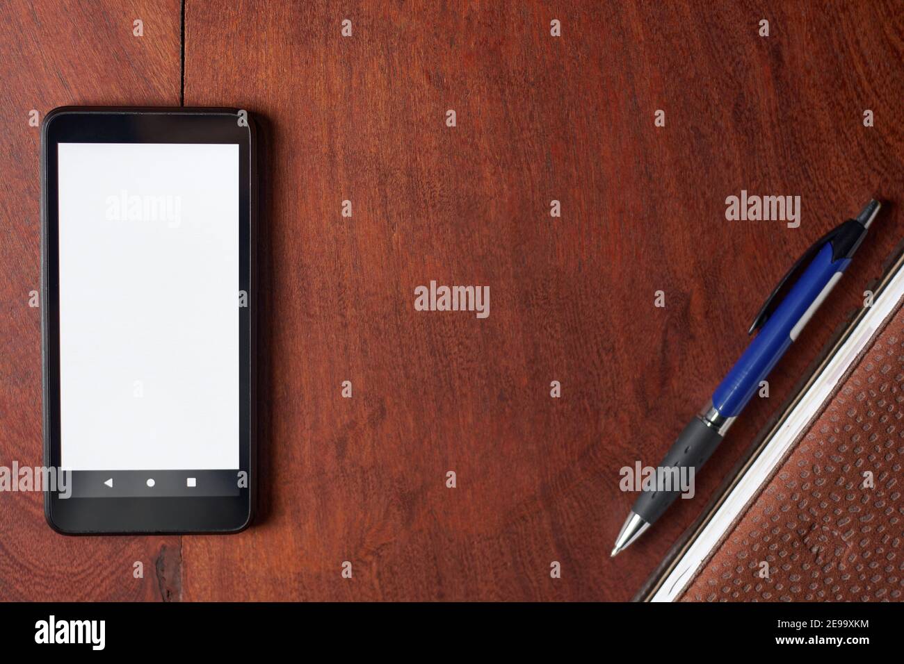 Black mobile phone with blank white screen, pen and notebook on rustic wooden table. Minimalistic top view of office setup with space for text. Work f Stock Photo