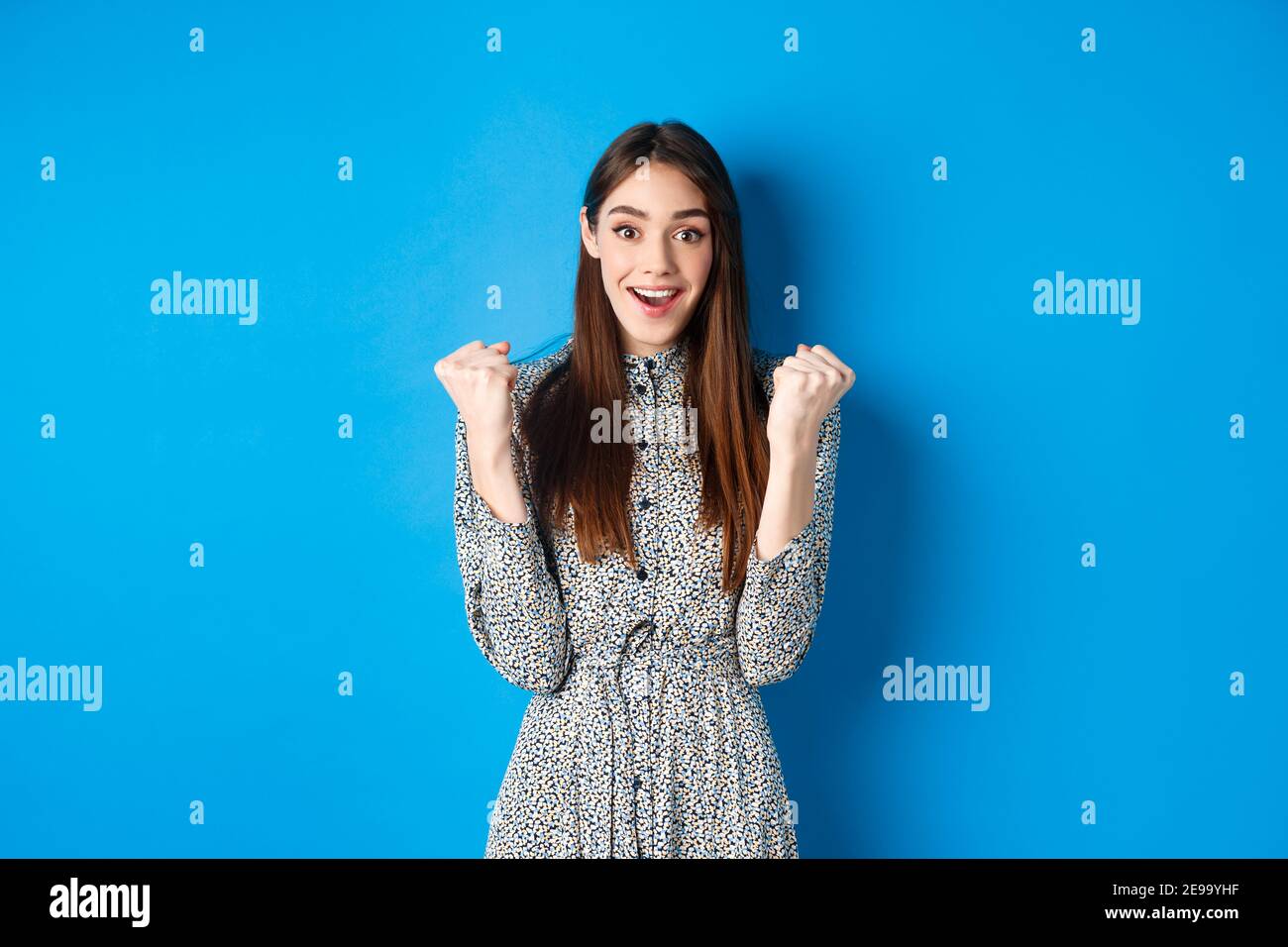 Excited woman looking relieved and hopeful, making fist pump and smiling, winning prize, celebrating achievement or victory, standing on blue Stock Photo