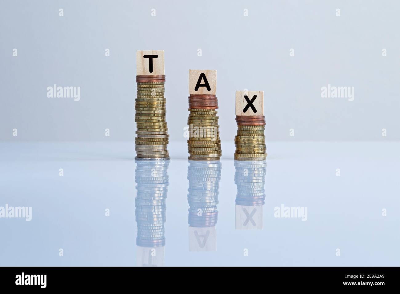 Word 'TAX' on wooden blocks on top of descending stacks of coins against gray background. Concept photo of tax cut, economy, business and finance. Stock Photo
