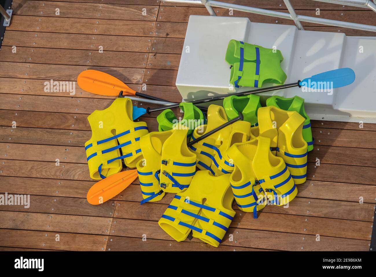 A pile of yellow and green life vests with boat paddles piled on a wooden dock and some white plastic stairs Stock Photo