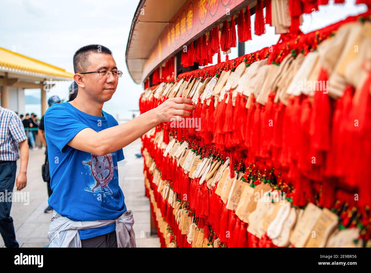 Chinese man is looking at wooden planks for writing prayers. Chinese red and wooden plates for wishes. Buddhist center Nanshan. Asian traditions. Stock Photo