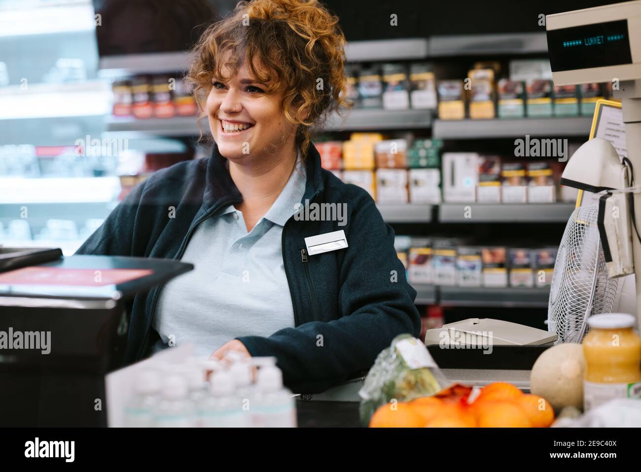 Happy woman cashier at a supermarket attending customer. Woman working at grocery store checkout talking with customer and smiling. Stock Photo