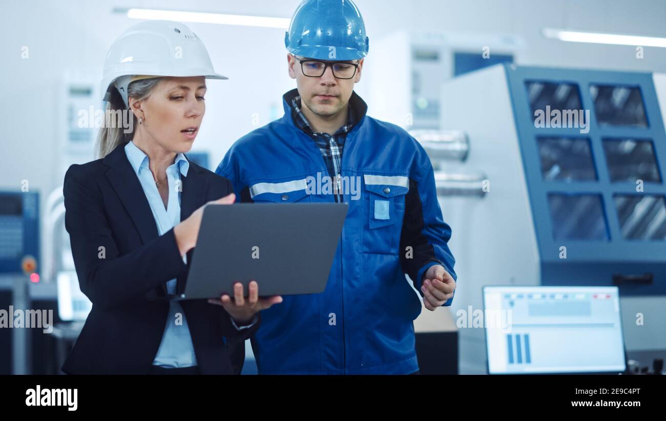 Female Manager and Project Engineer Wearing Hardhats Use Laptop in Industrial Factory, Talk, Plan Productivity Optimization. Production Line Workers Stock Photo