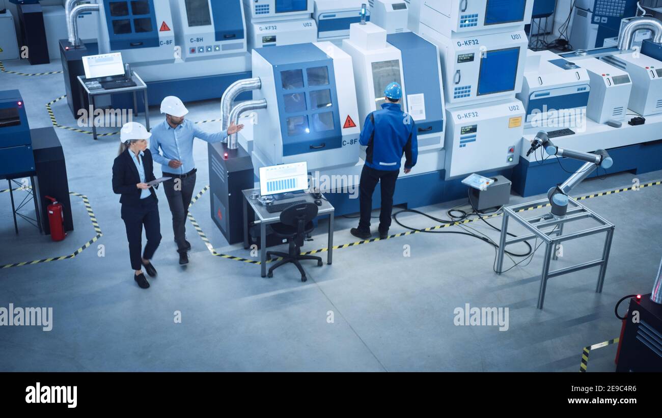 Industry Four Factory: Female Executive and Chief Engineer Inspect Facility Workshop, Look at robot arm working on Assembly Line. High-End CNC Stock Photo