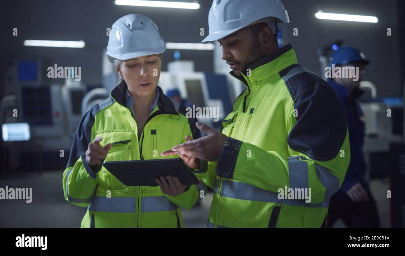Chief Engineer and Project Manager Wearing Safety Vests and Hard Hats, Use Digital Tablet Computer in Modern Factory, Talking, Optimizing Production Stock Photo