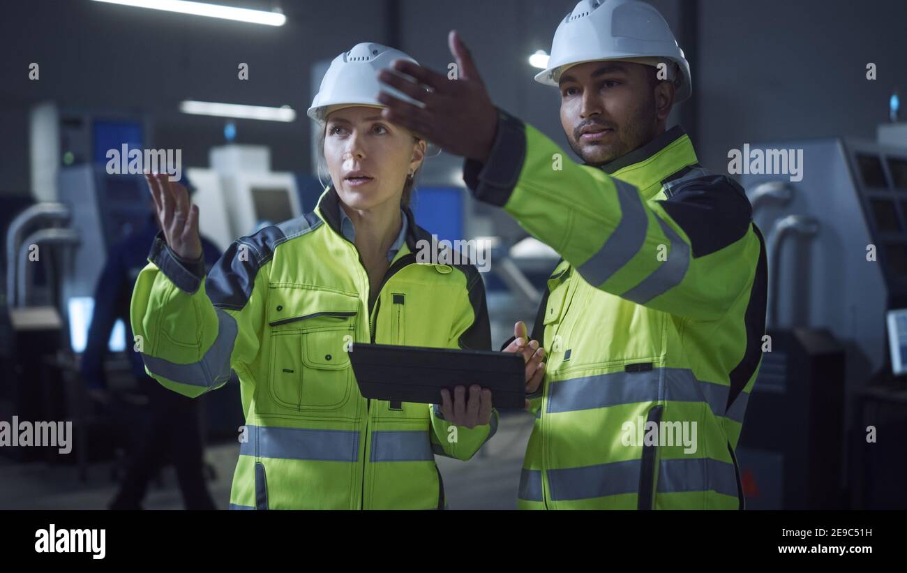 Chief Engineer and Project Manager Wearing Safety Vests and Hard Hats, Use Digital Tablet Computer in Modern Factory, Talking, Optimizing Production Stock Photo