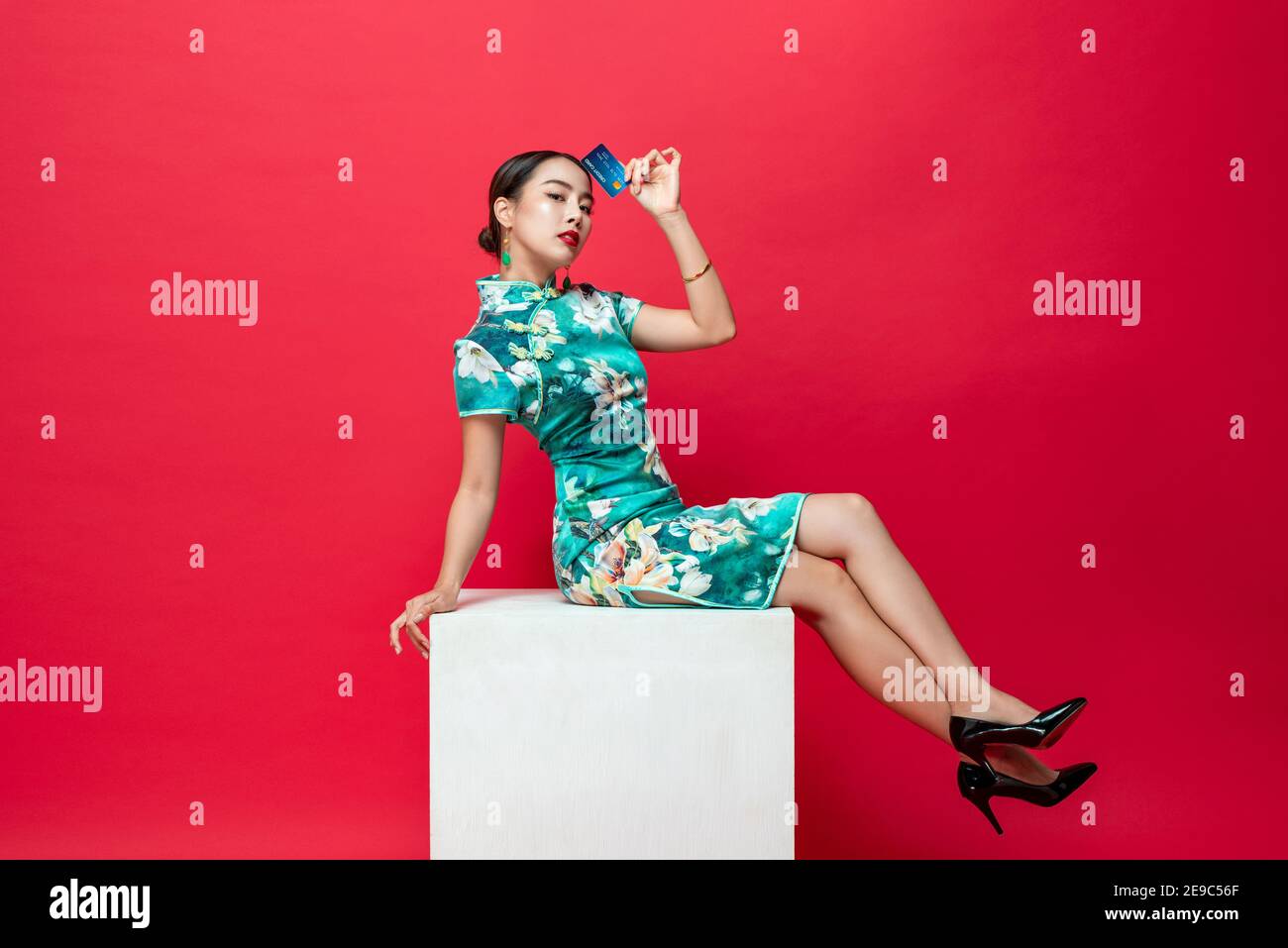 Beautiful Asian woman wearing oriental qipao dress showing credit card in hand sitting on stool in red studio background for Chinese new year shopping Stock Photo