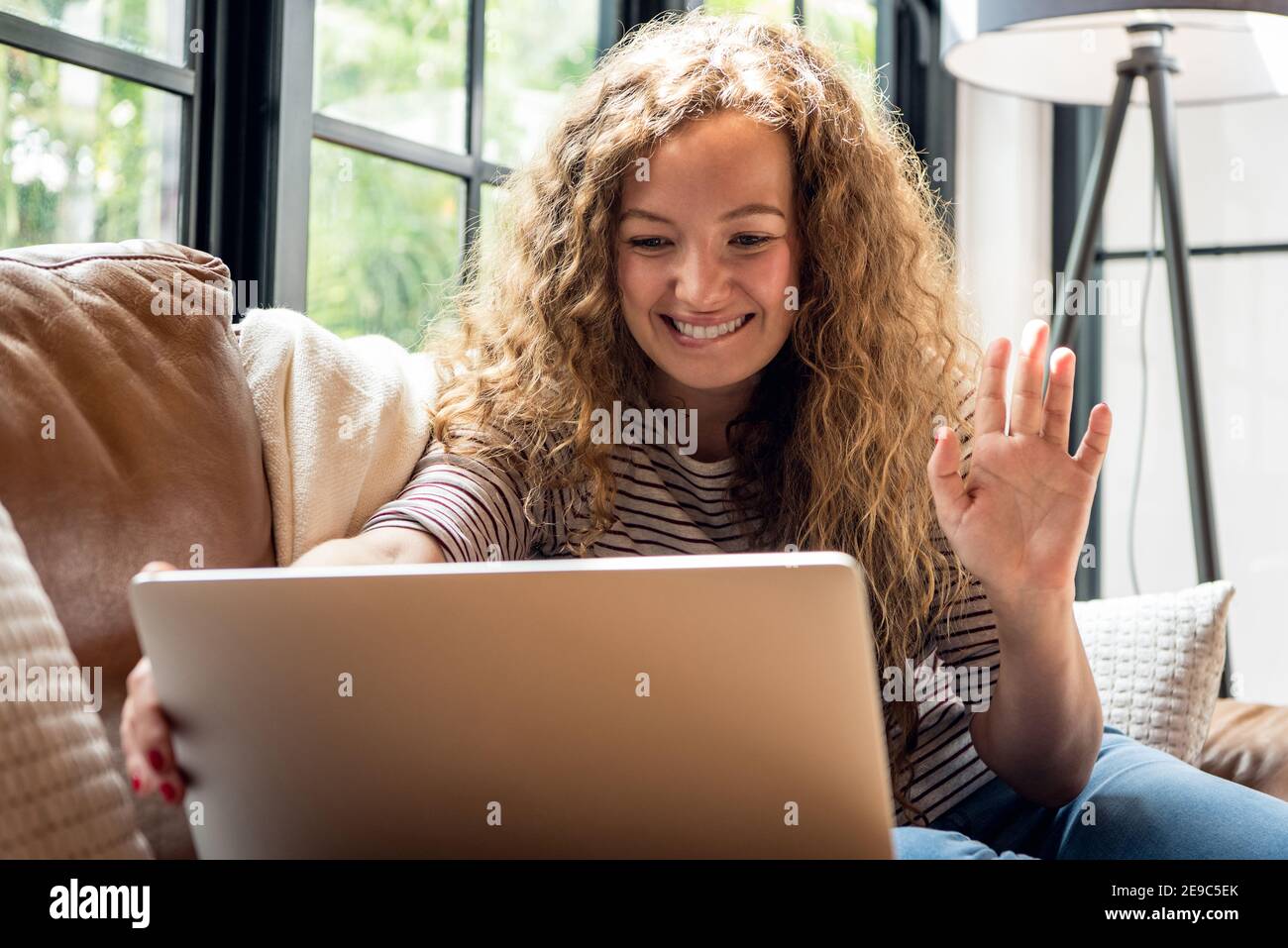 Pretty caucasian woman smiling and waving hand to the camera while making video call with laptop computer on the couch in living room at home Stock Photo