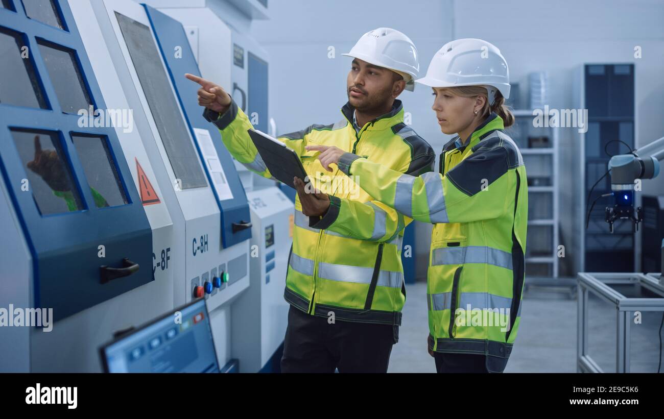 Chief Engineer and Project Manager Wearing Safety Vests and Hard Hats, Use Digital Tablet Controller in Modern Factory, Talking, Optimizing CNC Stock Photo