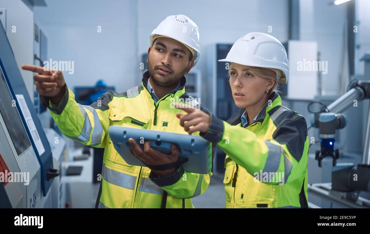 Chief Engineer and Project Manager Wearing Safety Vests and Hard Hats, Use Digital Tablet Controller in Modern Factory, Talking, Optimizing CNC Stock Photo