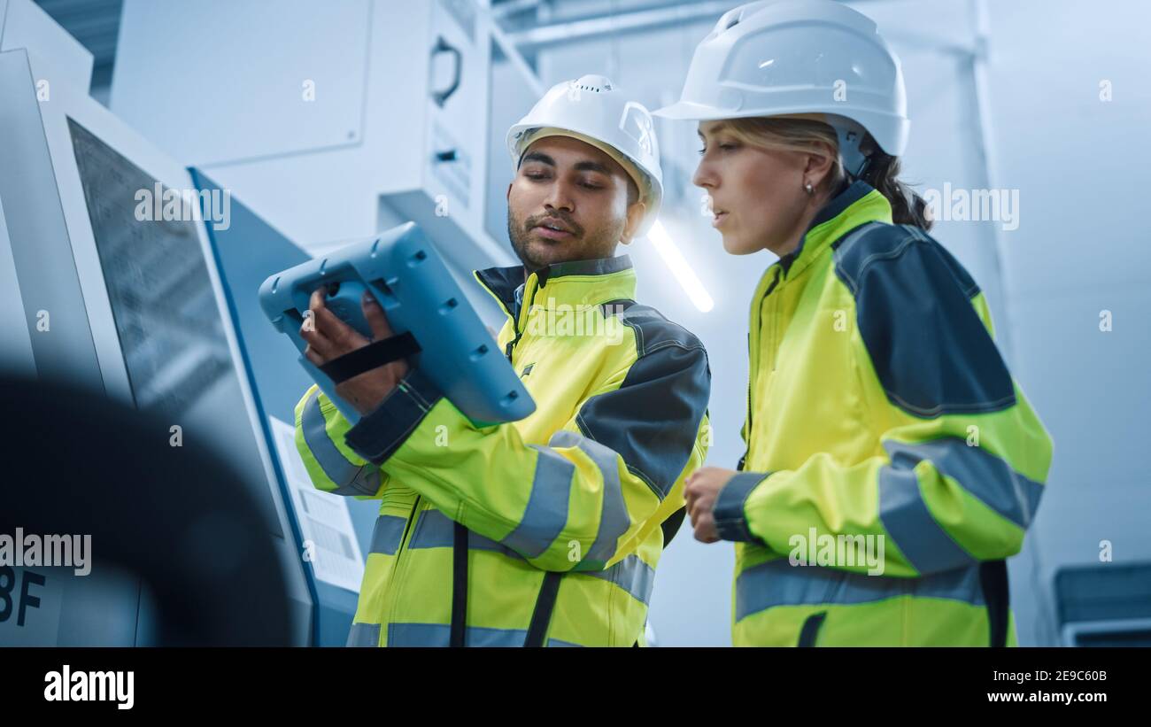 Chief Engineer and Project Manager Wearing Safety Vests and Hard Hats, Use Digital Tablet Controller in Modern Factory, Talking, Programming Machine Stock Photo