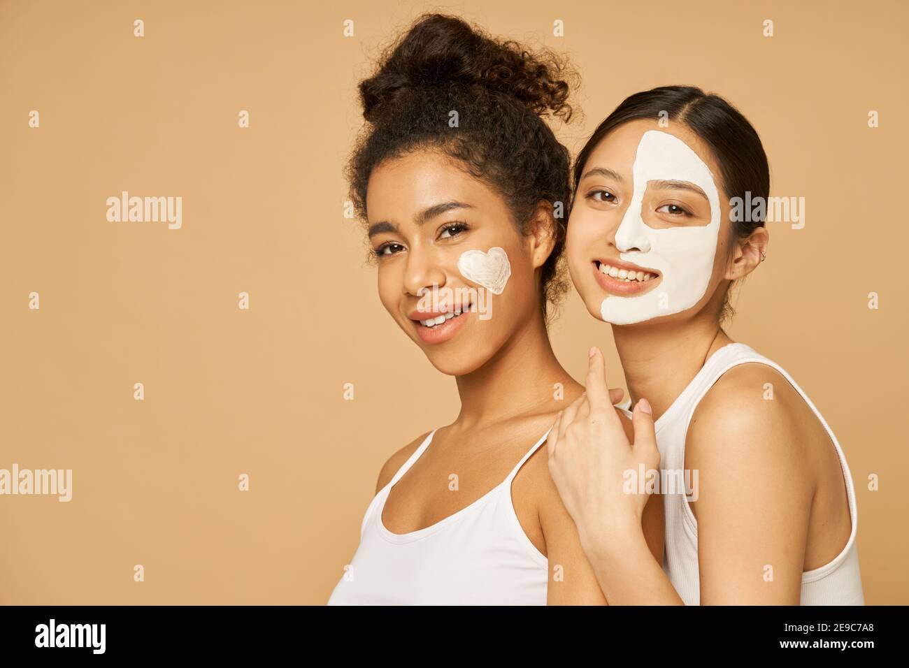 Two young female friends having fun, smiling at camera while posing with facial masks on isolated over beige background. Skincare, beauty concept Stock Photo