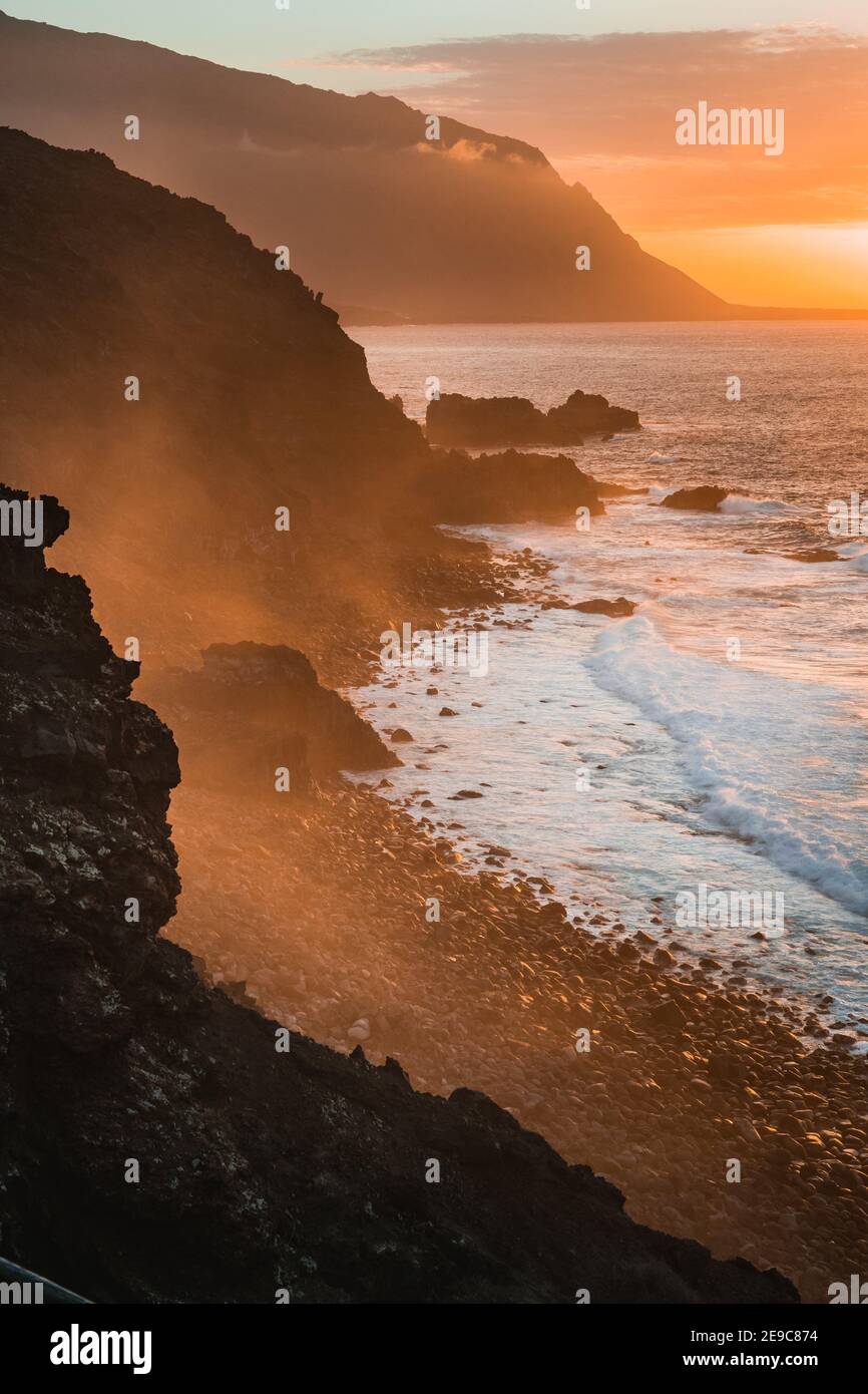 Waves hit rocky coastline at sunset, beautiful layered cliffs with mist rolling in from ocean. Atmospheric view sightseeing. Stock Photo