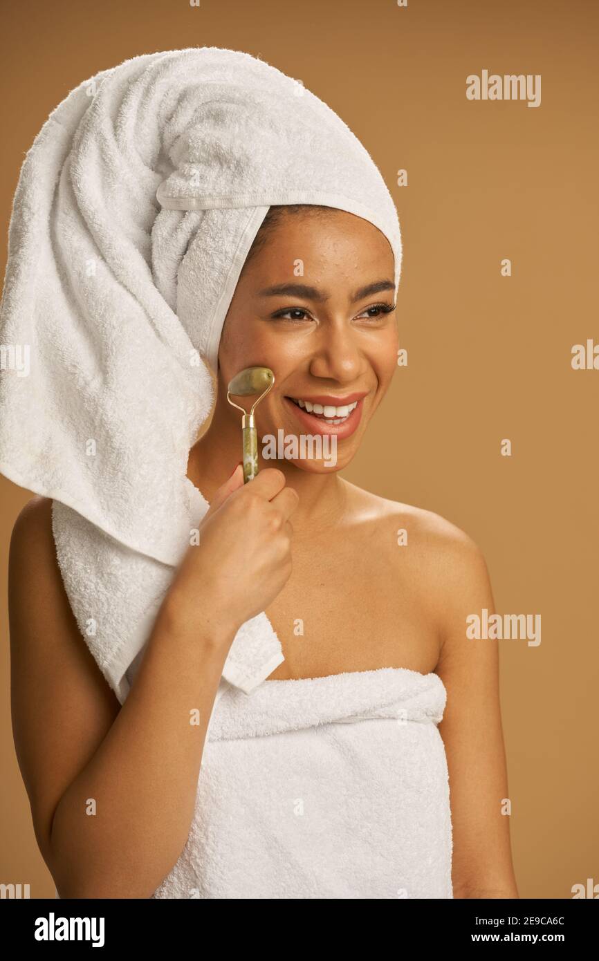 Cheerful mixed race young woman after shower smiling aside while using jade roller for massaging