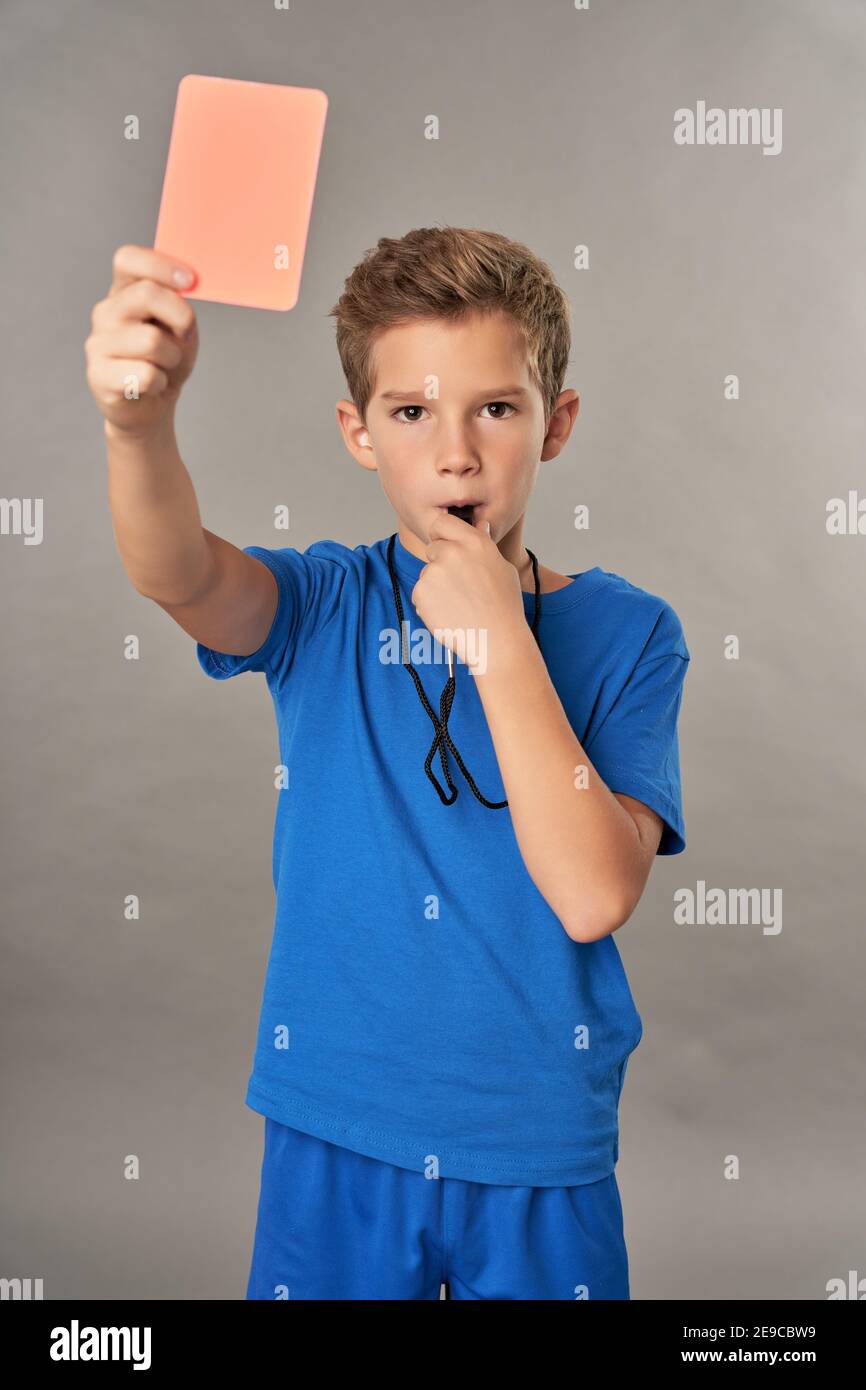 Adorable male kid using referee whistle and showing penalty card while standing against gray background Stock Photo