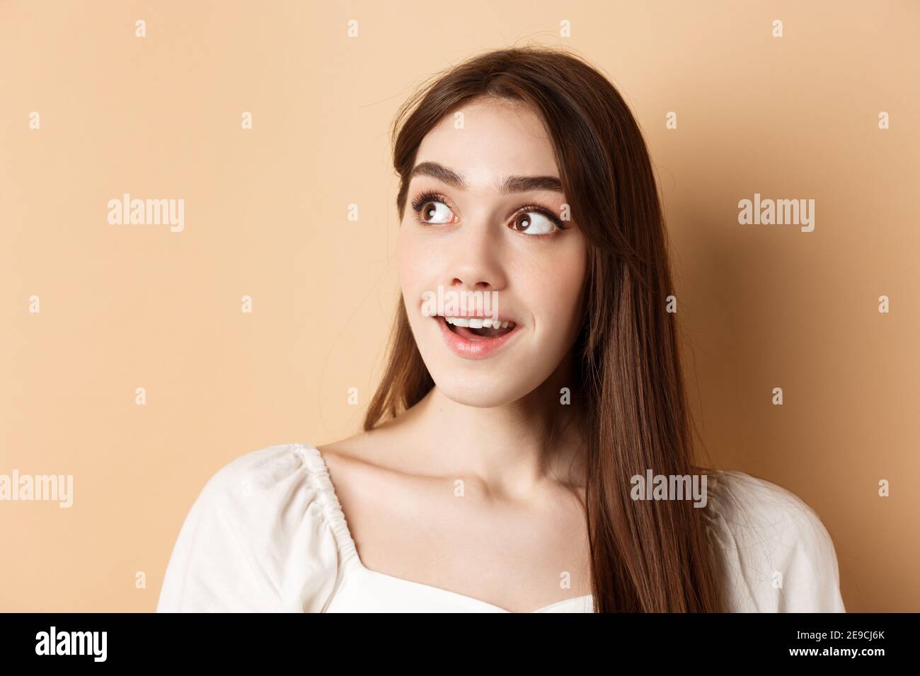 Dreamy beautiful girl looking at upper left corner logo, checking out banner, standing thoughtful on beige background Stock Photo