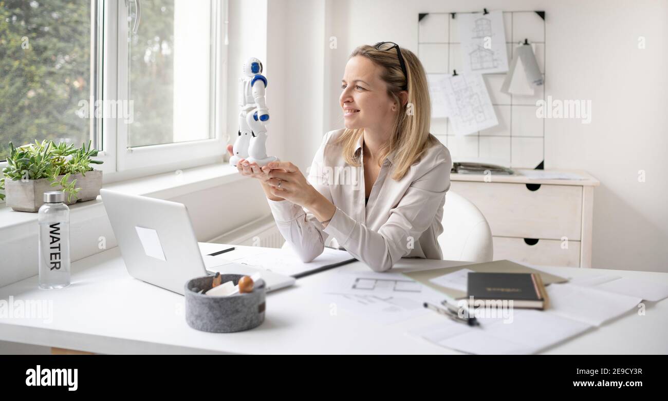pretty blonde business woman is sitting in her office and is working with her new little robot Stock Photo