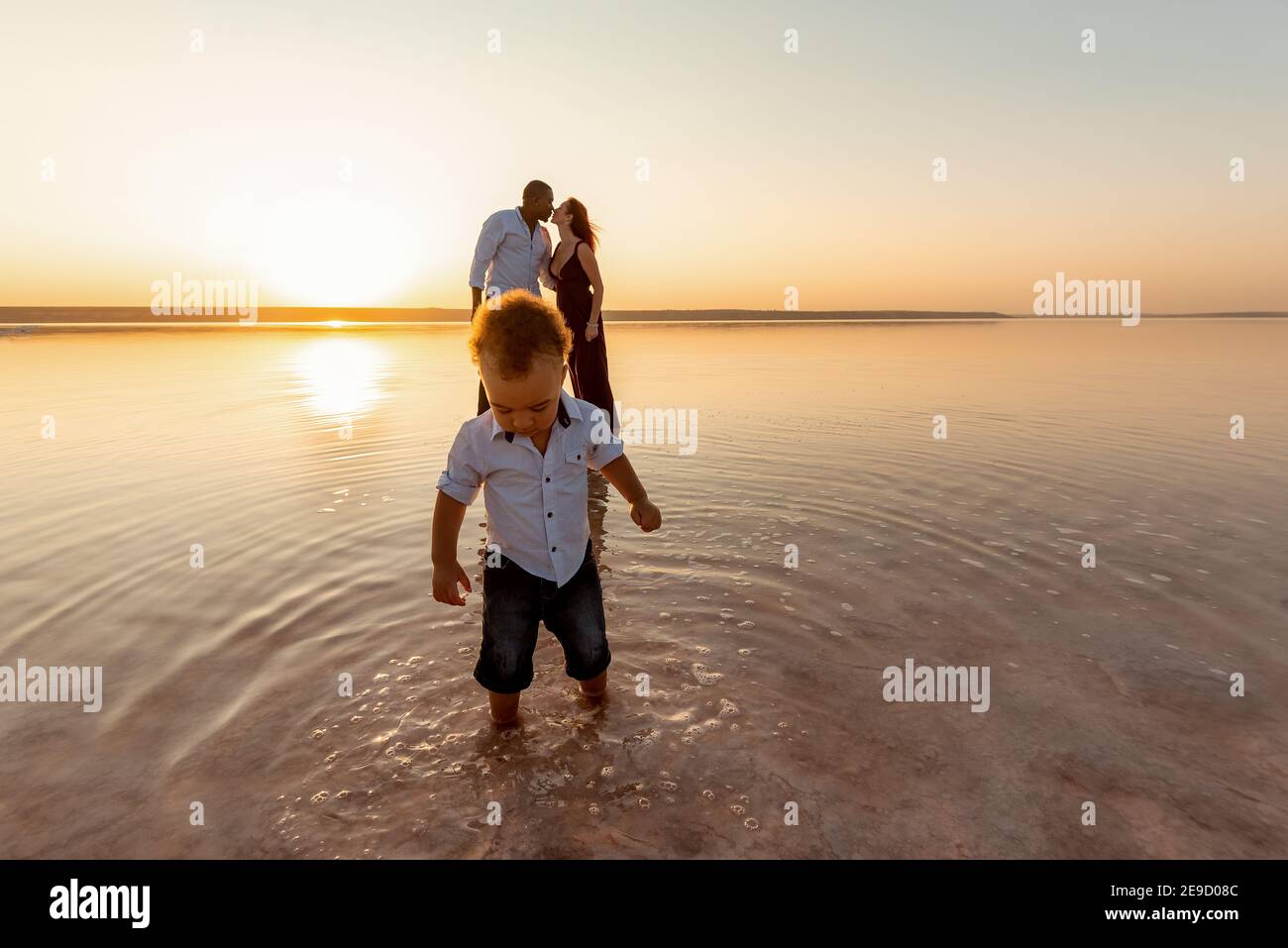 Portrait of little mixed race boy. Kissing parents on background. Happy multi ethnic family at the beach. Beautiful sunset Stock Photo