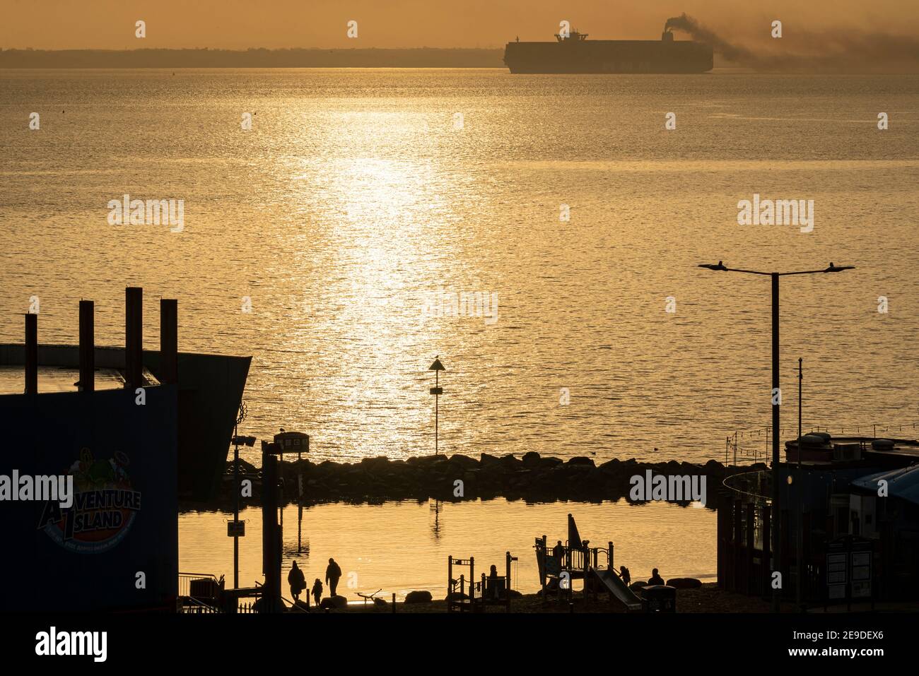 HMM Le Havre container ship on the Thames Estuary late afternoon off Southend on Sea, Essex, UK, belching exhaust. Children playing near lagoon. Dusk Stock Photo