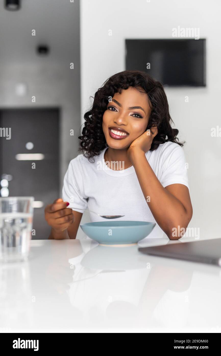 Young healthy laughing african woman eating breakfast and fruits in kitchen in the morning Stock Photo