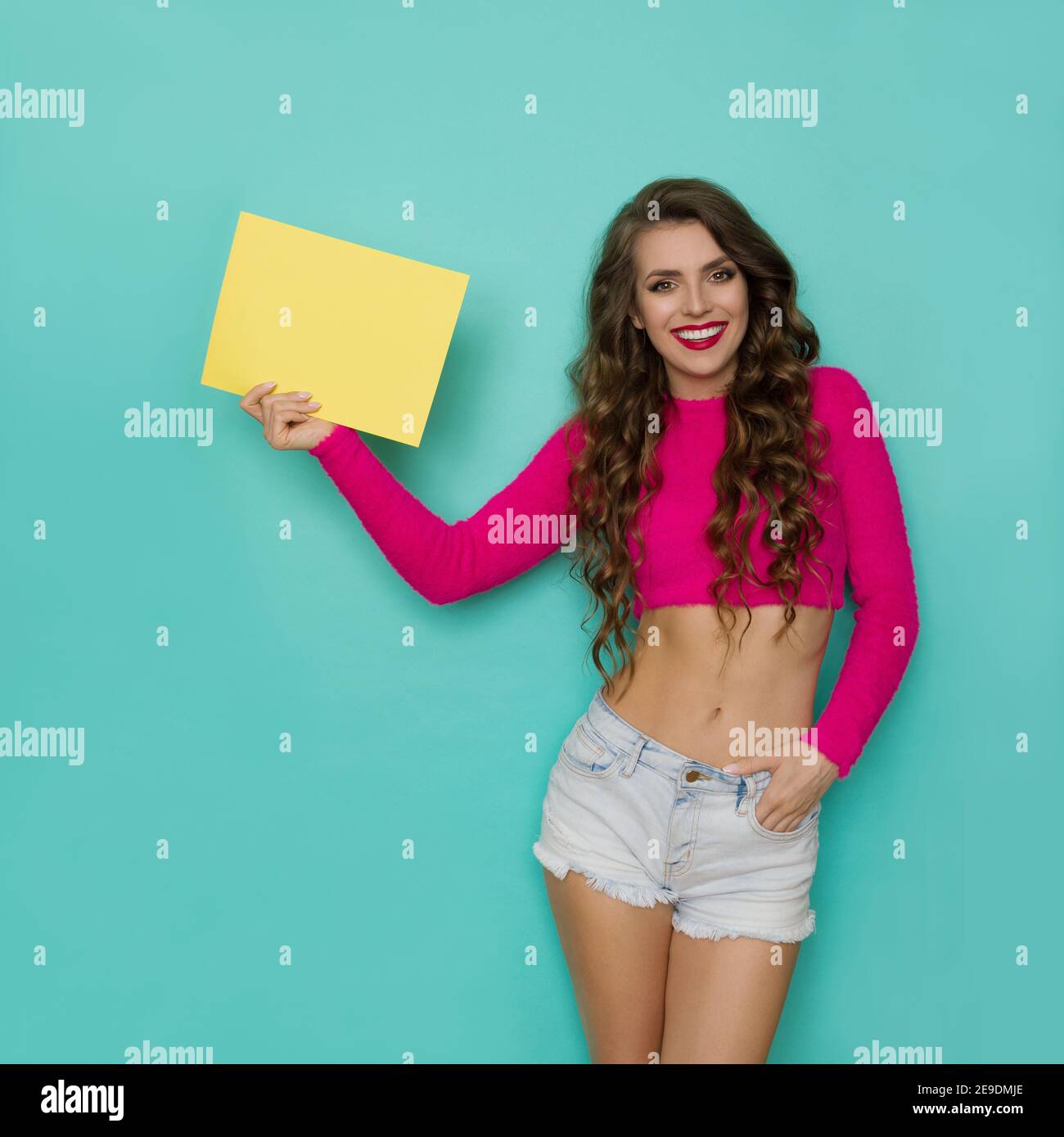 Beautiful young woman is standing relaxed with hand in pocket, showig yellow sheet of paper and smiling. Front view. Three quarter length studio shot Stock Photo
