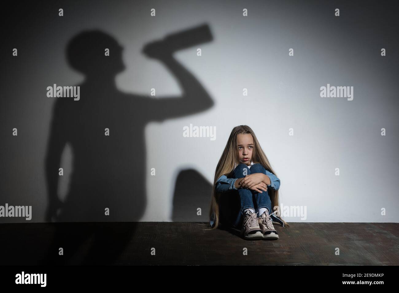 Domestic physical violence, abusing. Scared little caucasian girl, victim sitting close to white wall with shadow of angry threatening father with alcohol addiction. Awareness of social problem. Stock Photo