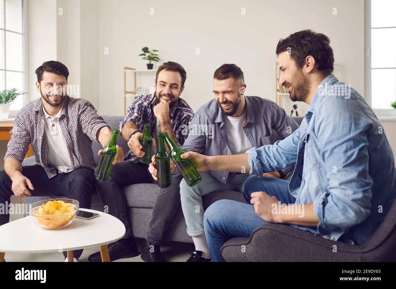 Group of friends sitting on couch, laughing and clinking beers after funny toast Stock Photo