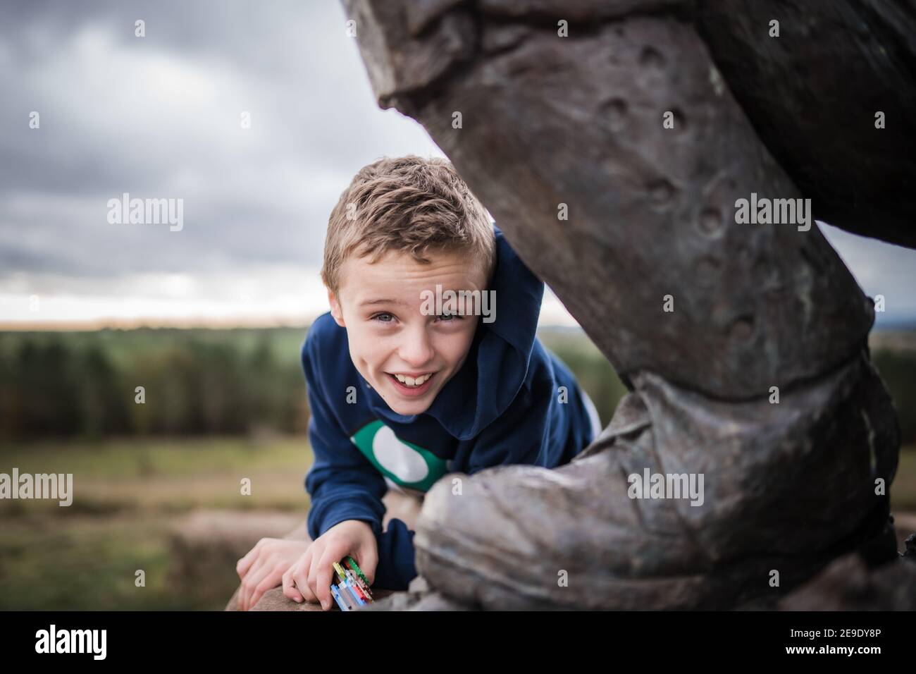 Happy smiling laughing young boy exploring outdoors forest wearing blue hooded top behind giant foot of bronze statue Stock Photo
