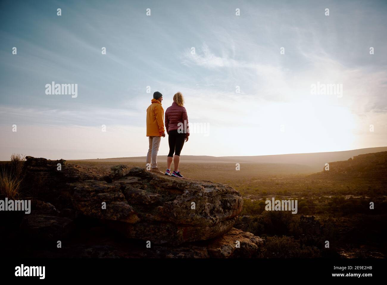 Rear view of young man and woman standing looking at beautiful sunrise on mountain cliff peak Stock Photo
