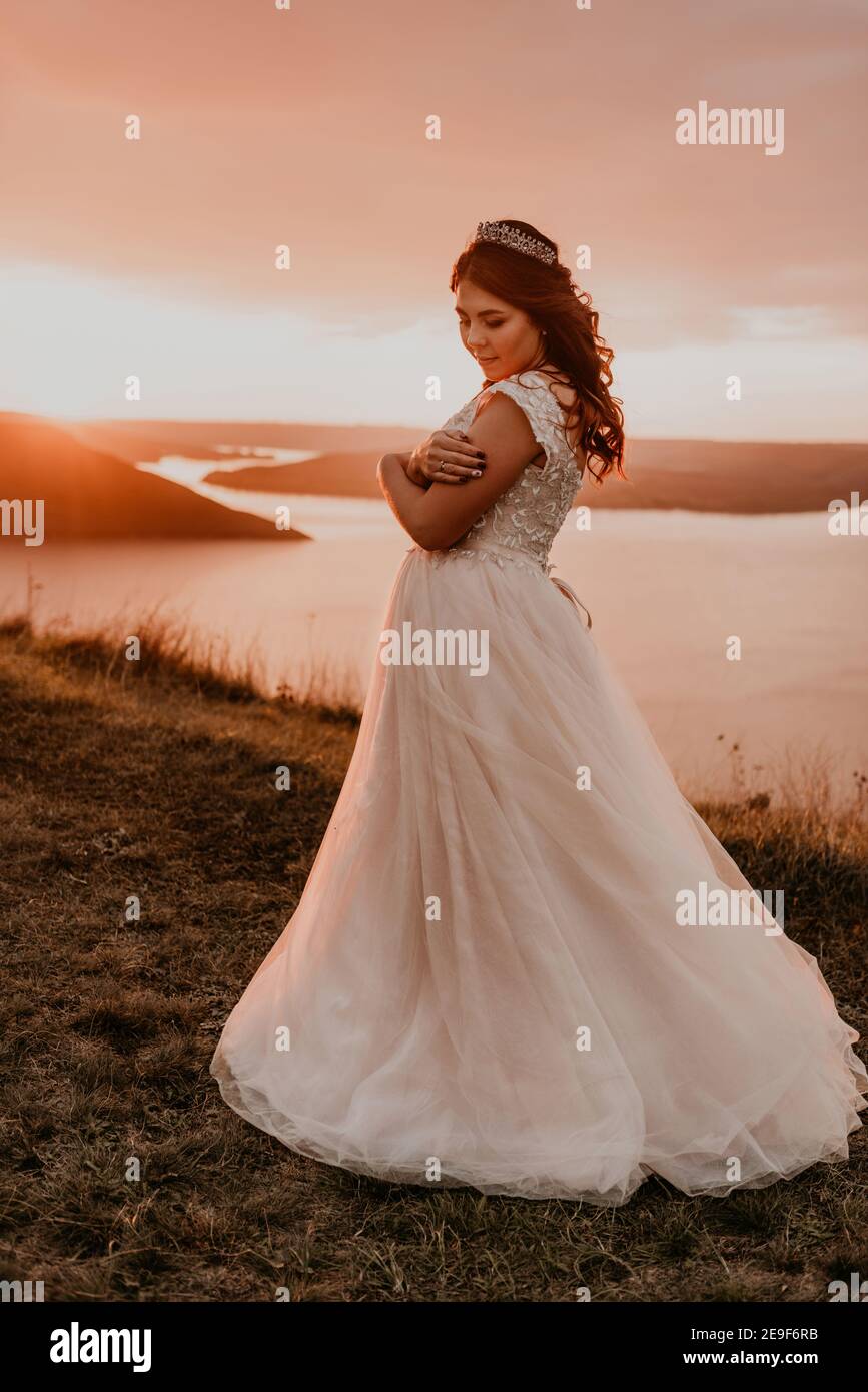 young brunette bride in white wedding dress with a crown on her head stands on cliff against the background of the river and islands After sunset Stock Photo