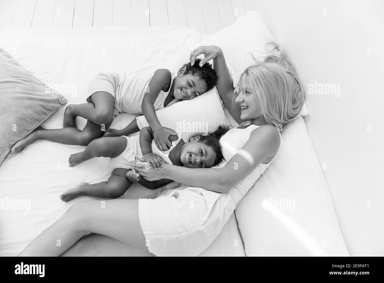 Young Caucasian blond mother plays, lies on white bed with African American daughter son. Happy multi ethnic family morning. Woman hugs, strokes child Stock Photo