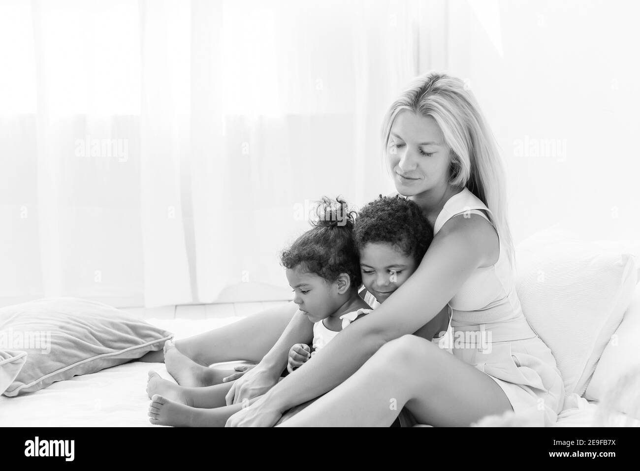 Young Caucasian blond mother plays, lies on white bed with African American daughter son. Happy multi ethnic family morning. Woman hugs, strokes child Stock Photo