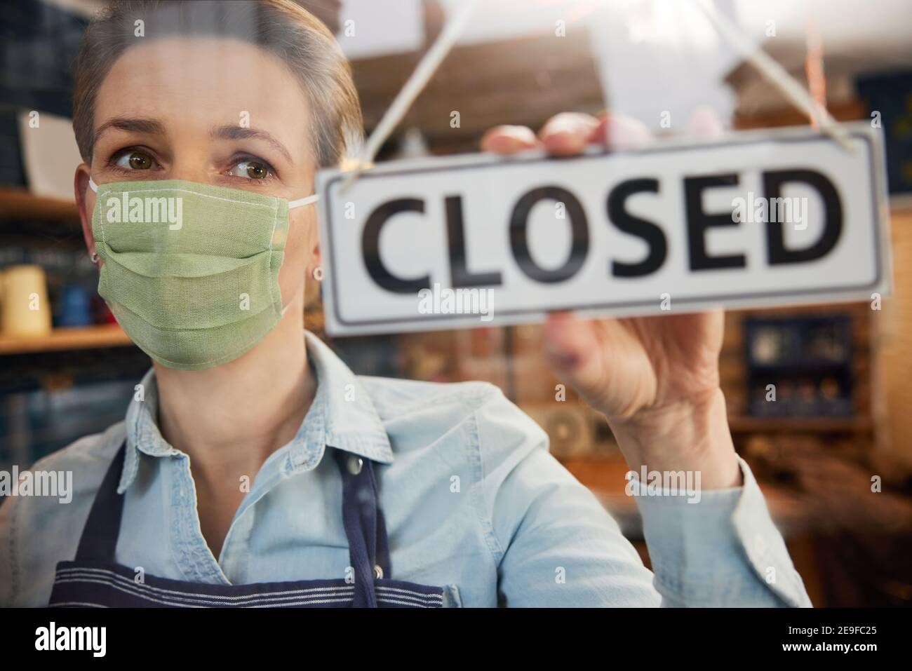 Female Owner Of Small Business Wearing Face Mask Turning Round Closed Sign During Health Pandemic Stock Photo