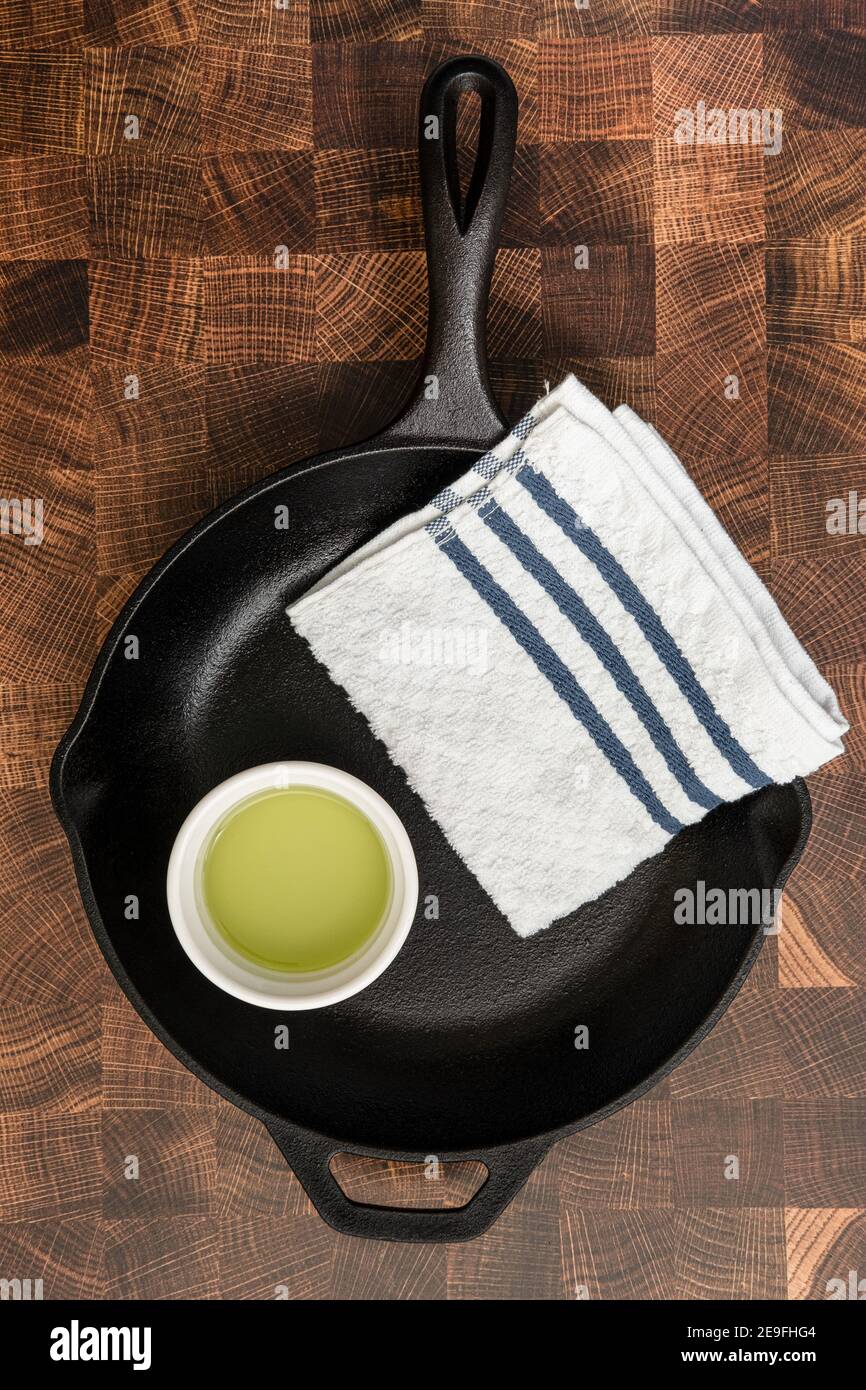 A cast-iron skillet being prepared to be seasoned, with a cup of grapeseed oil and a towel on a cutting board board Stock Photo