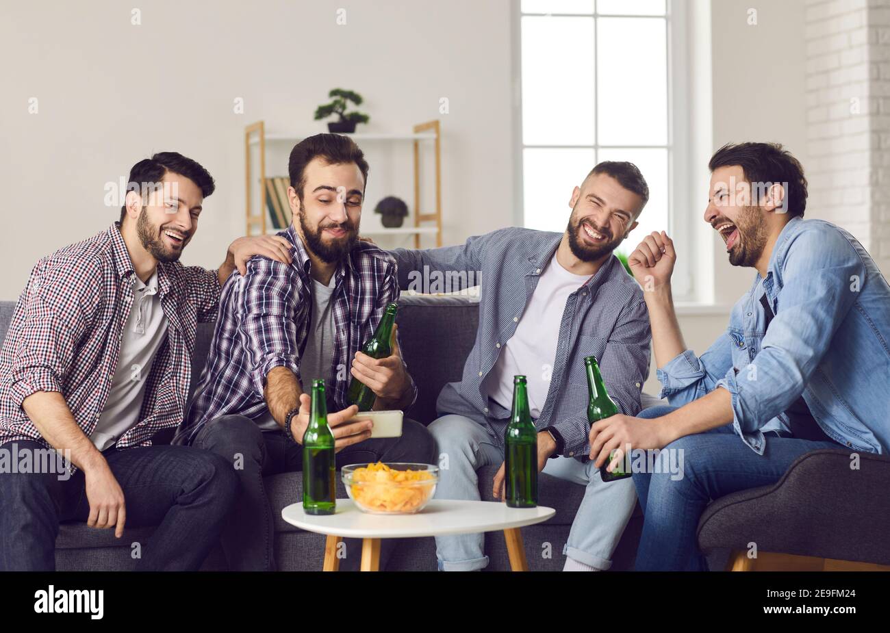 Smiling excited men friends drinking beer, eating snacks and making selfie during home party Stock Photo