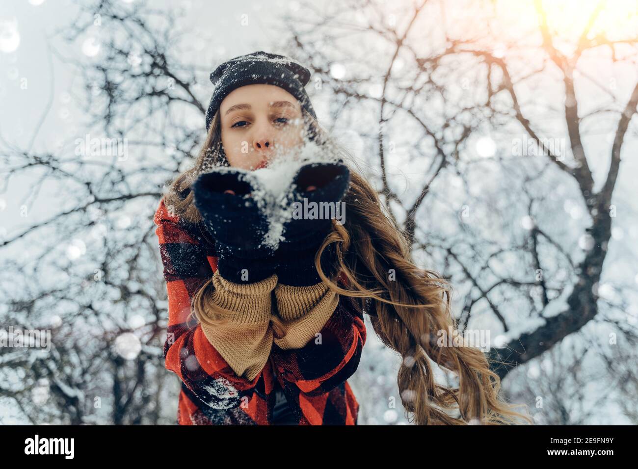 Woman Wearing Warm Winter Clothes And Hat Blowing Snow In Winter Park. Flying Snowflakes. Sunny day. Joyful Beauty young girl Having Fun in frosty Stock Photo