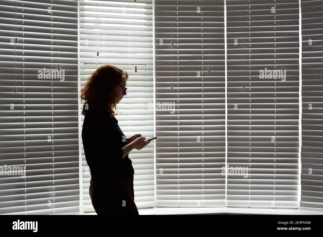 Photograph of a young woman in shilouette standing in front of a large window during UK Covid 19 isolation. Woman is holding a mobile phone. Stock Photo
