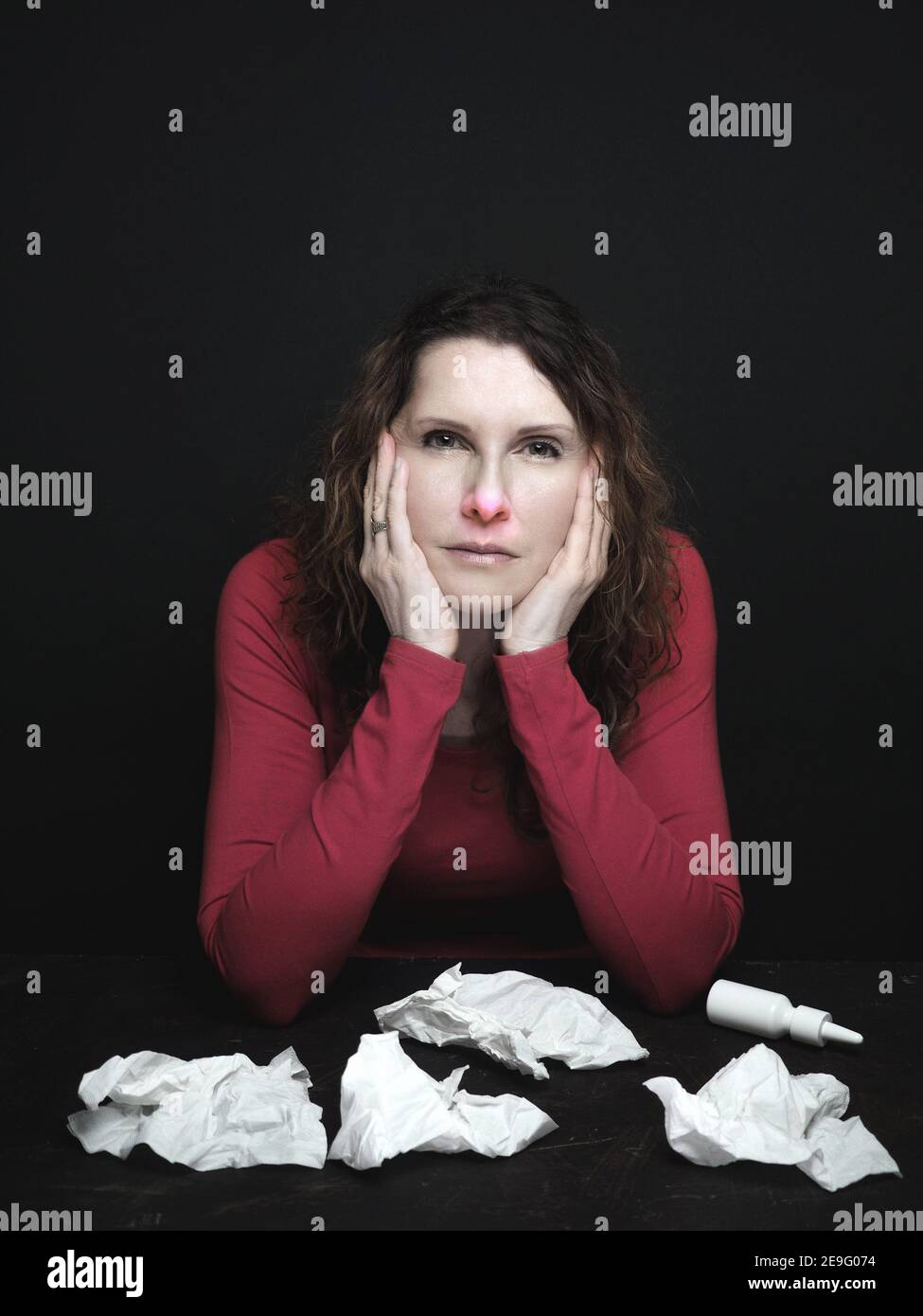 Caucasian woman with stuffy red nose and shiny eyes suffering from cold. Used paper tissues and nasal spray on the table. Copy space.  Stock Photo