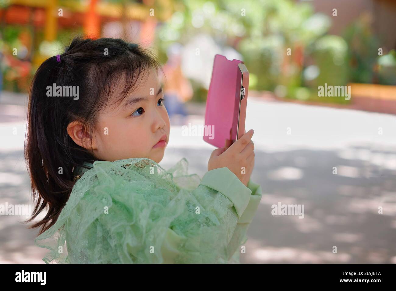 A cute young Asian girl is holding a pink smartphone, trying to take a picture on her vacation. Stock Photo