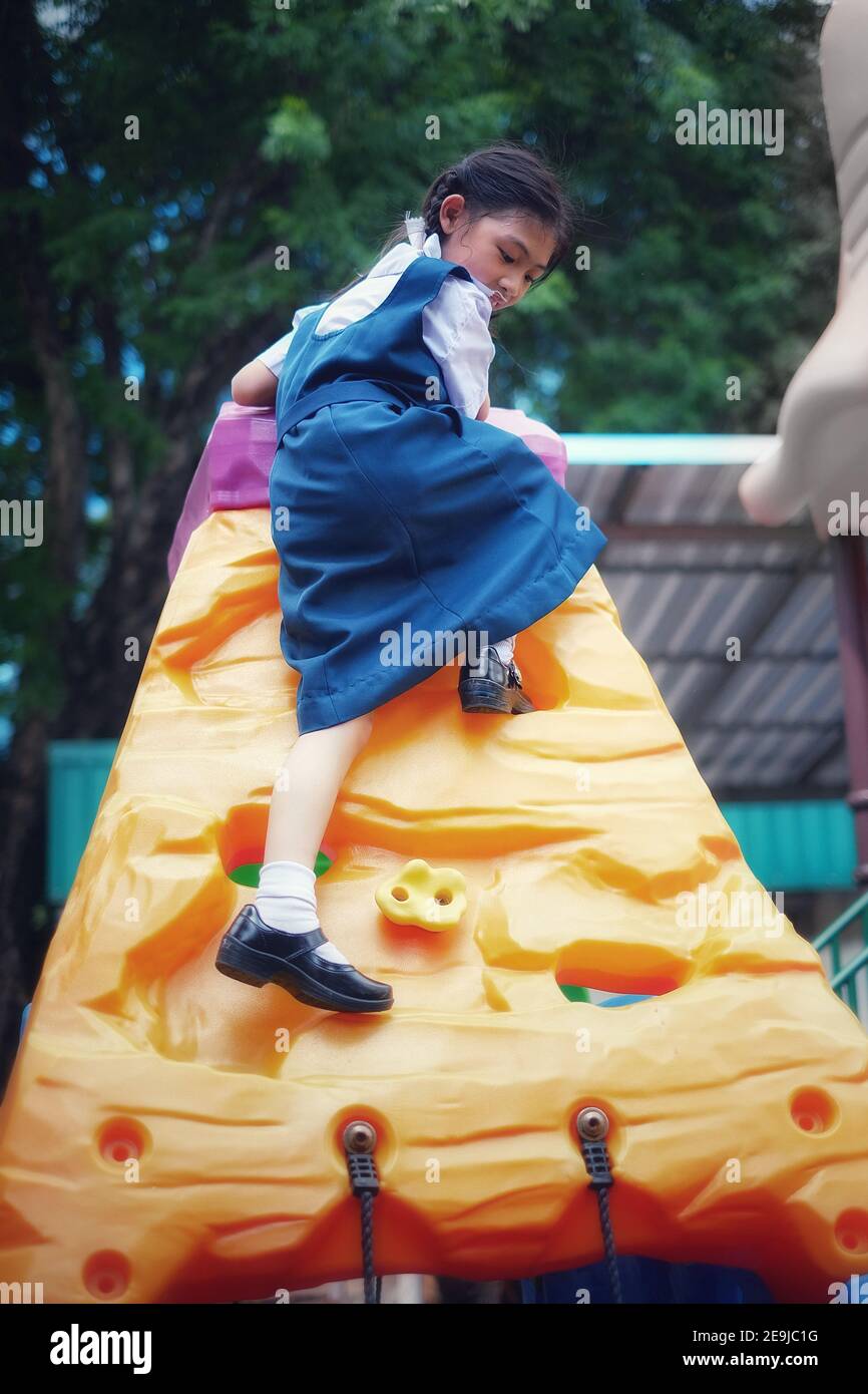 A cute young Asian girl in white and blue school uniform is playing rock climbing at a playground, exercising and having fun. Stock Photo