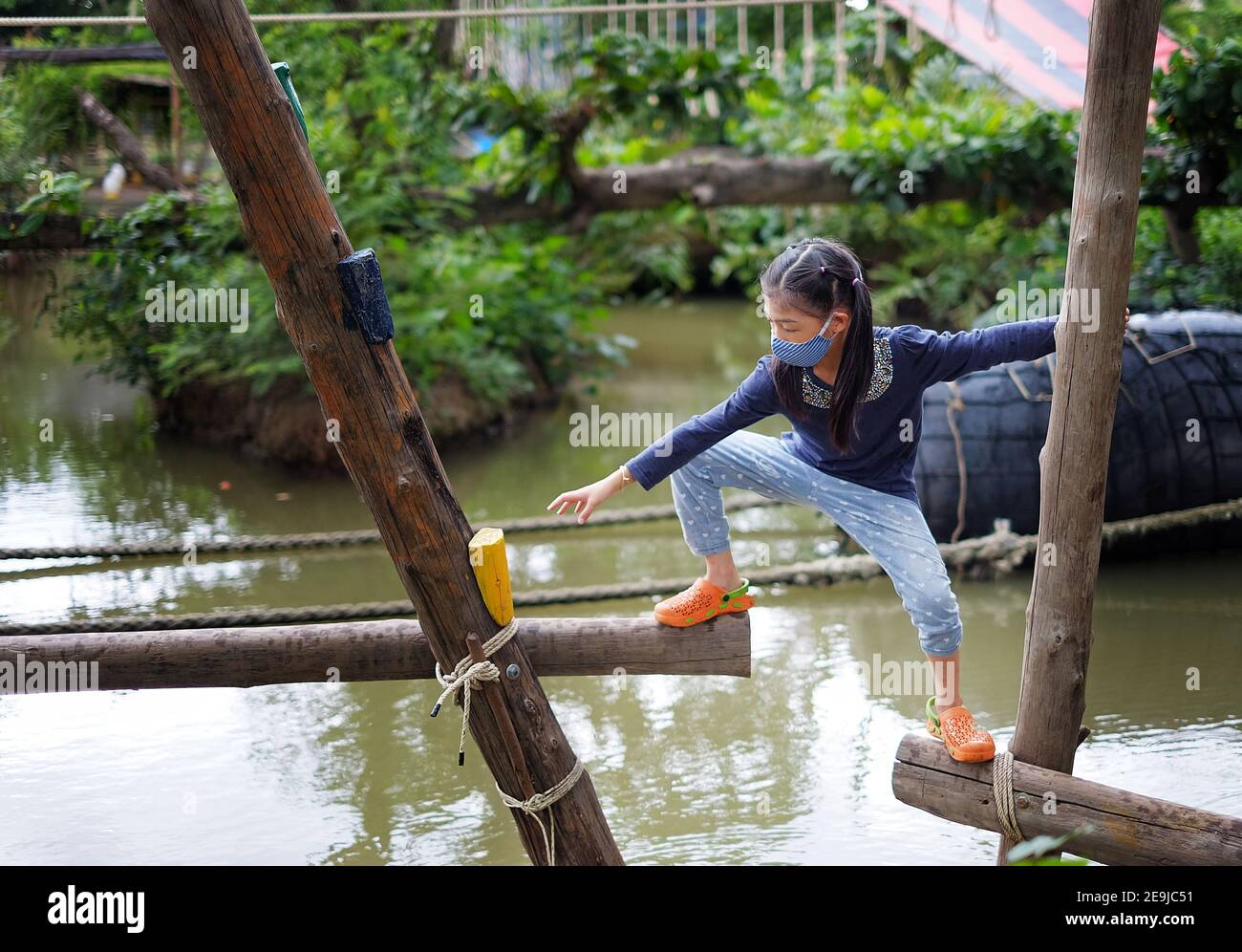 A cute young Asian girl trying to cross a small canal through a timber structure with opening gaps, being brave but cautious to reach her destination. Stock Photo