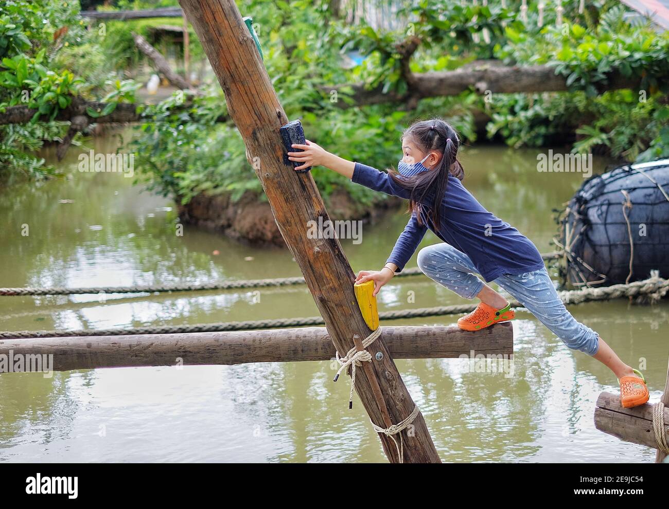 A cute young Asian girl trying to cross a small canal through a timber structure with opening gaps, being brave but cautious to reach her destination. Stock Photo