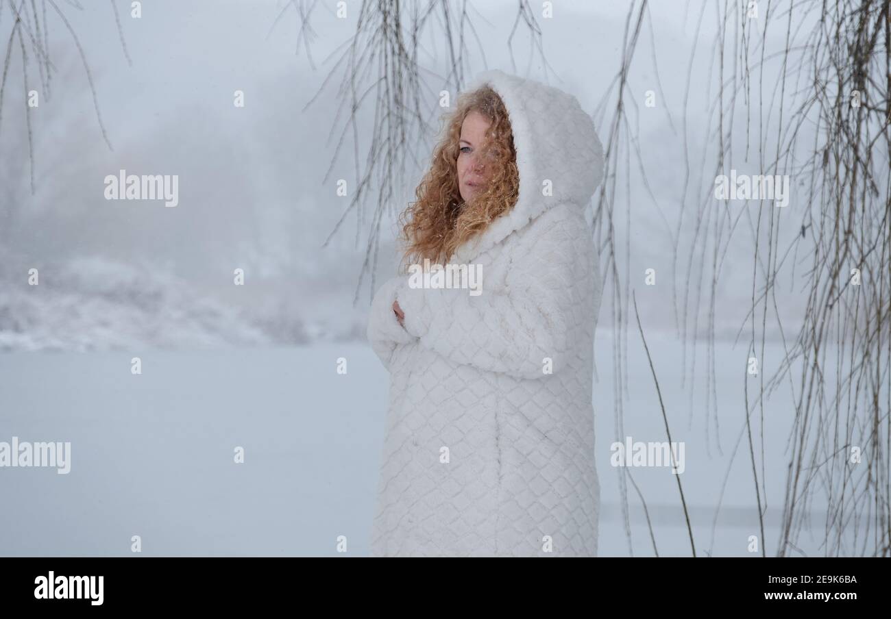 redhead mature woman in her fifties with red curly hair and white hooded coat freezes in the snowy winter lake landscape Stock Photo