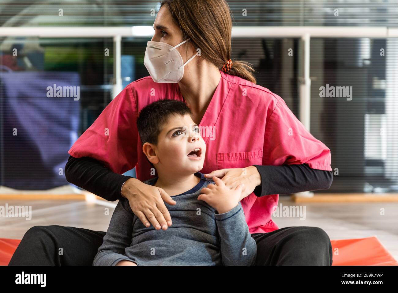 disabled child and physiotherapist on a red gymnastic mat doing exercises. pandemic mask protection Stock Photo