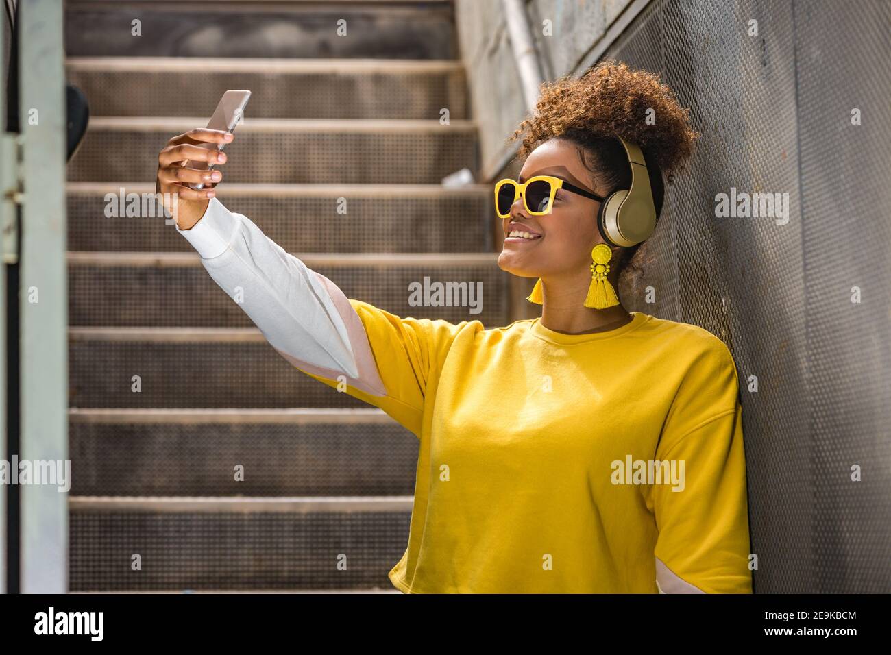 Happy millennial African American female in vivid yellow wear and trendy sunglasses taking selfie on mobile phone while listening to music with headph Stock Photo