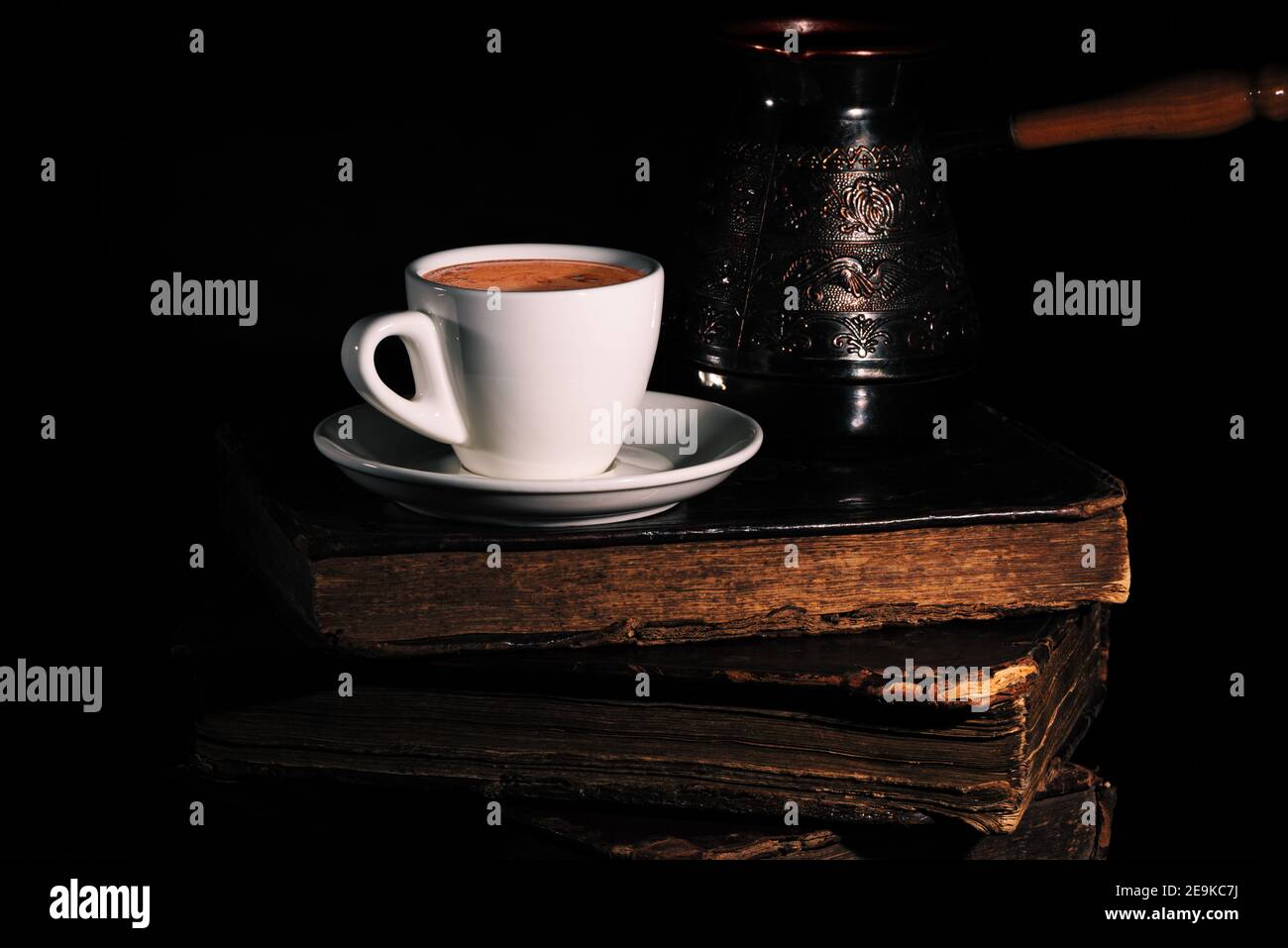 White cup of coffee and copper cezve on stack of old shabby books in the dark. Low key. Closeup Stock Photo