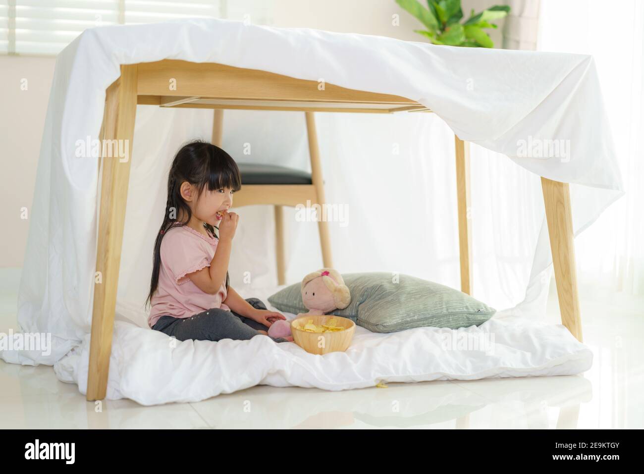 Asian cute little girl eating snack while sitting in a blanket fort in living room at home for perfect hideout away from their other family members an Stock Photo