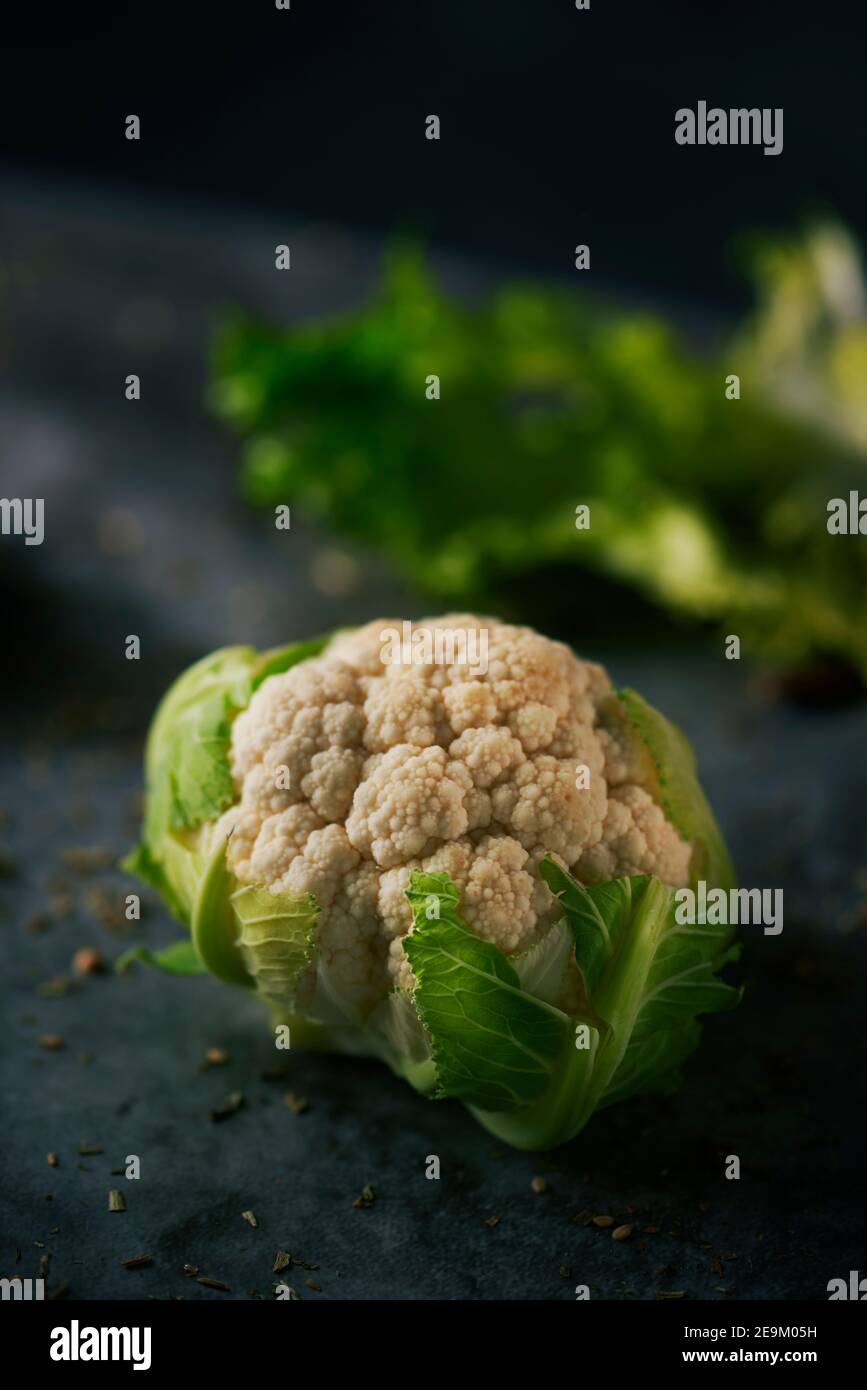 closeup of a raw baby cauliflower head placed on a dark stone surface sprinkled with different seeds and spices Stock Photo